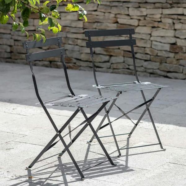 A pair of carbon black metal folding bistro chairs positioned on a light stone patio in front of a rustic stone wall.
