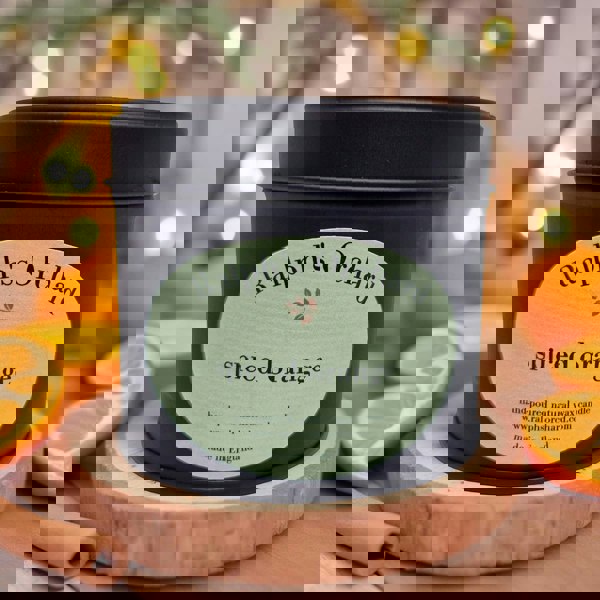 Candle in a black tin labeled 'Ralph's Orchard spiced orange' on a wooden coaster with oranges and cinnamon sticks.