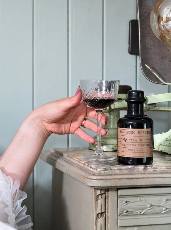 A hand holds a coupe glass containing a dark liquid next to a bottle of Espresso Martini mix by Prohibition Cocktails on a distressed wooden table, with a light paneled wall in the background.