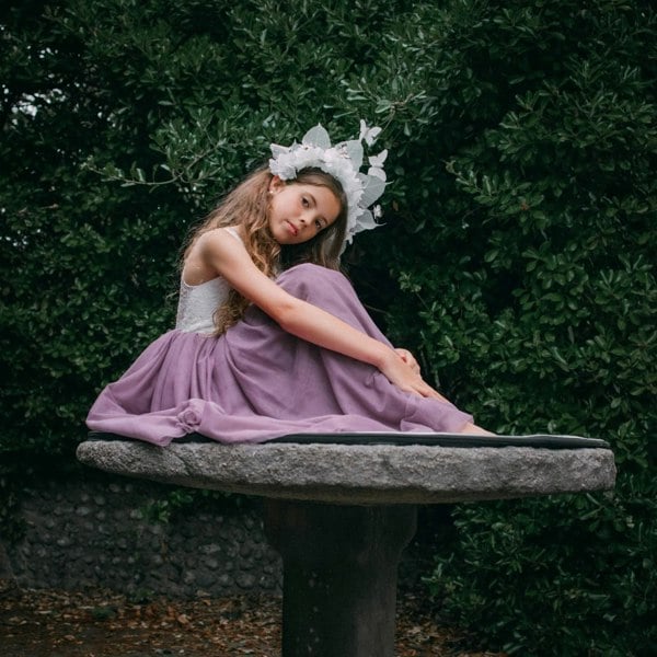 Young girl in a purple dress sitting on a stone bench with greenery in the background