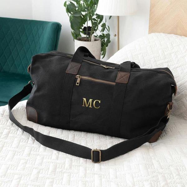 A personalised black waxed canvas holdall with gold initials MC on a white quilted bed runner.