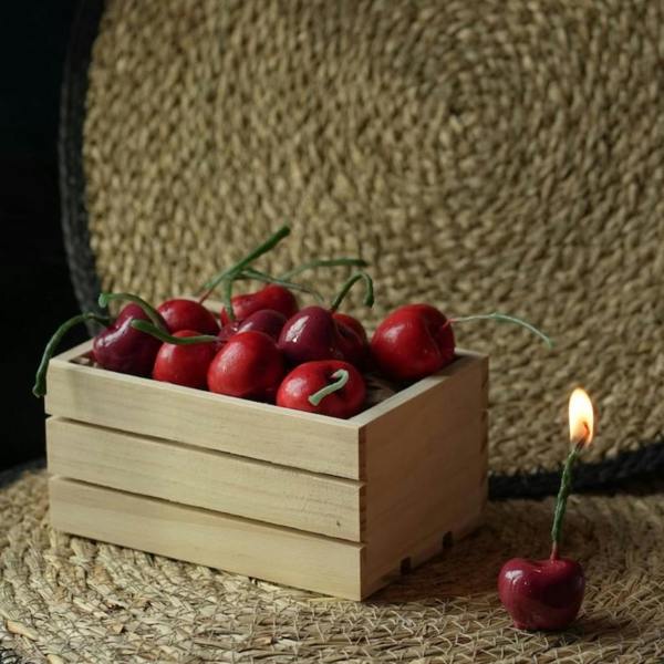 A small miniature wooden crate packed full of unlit red cherry-shaped candles, with one single lit cherry candle standing beside it on a woven placemat.