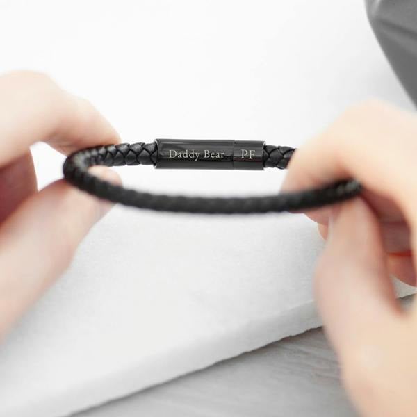 A close-up of a Personalised Men's Leather Bracelet held in two hands, showing a black woven leather band and an engraved clasp reading Daddy Bear PF.