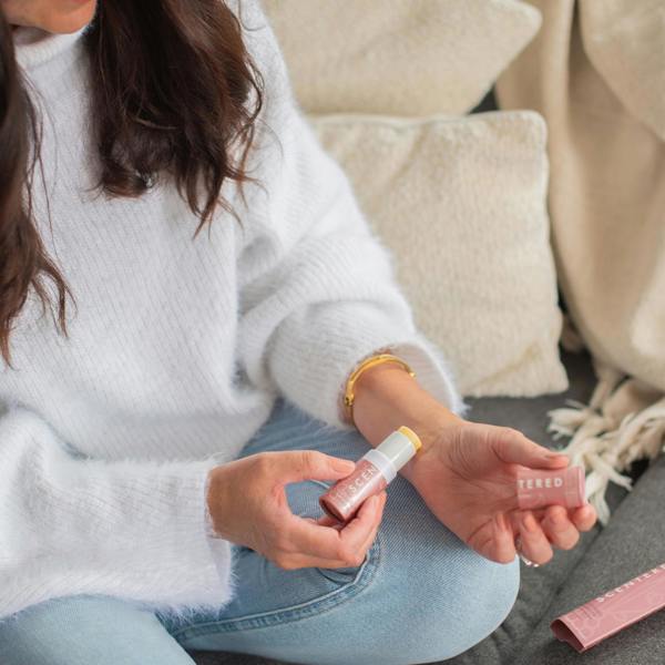 Woman sitting on a couch holding a balm and applying on her wrist.