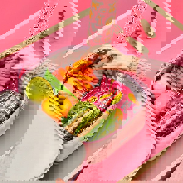 Lunchbox filled with green apple, orange, sandwich, granola bars, carrots, and Walkers crisps. A hand reaches for carrots. Nearby is a decorated water bottle on a pink table. The name "Amelia" is on the lunchbox.