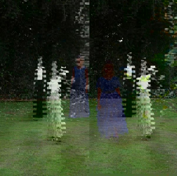 Two young girls in formal dresses standing on a grassy area with trees in the background.