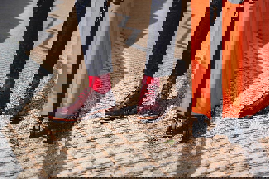 Person wearing brown shoes and red socks standing next to an orange suitcase on a paved walkway.