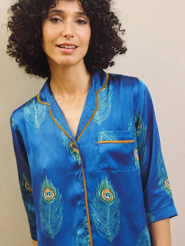 Woman wearing a blue satin pajama top with peacock feather patterns against a beige background