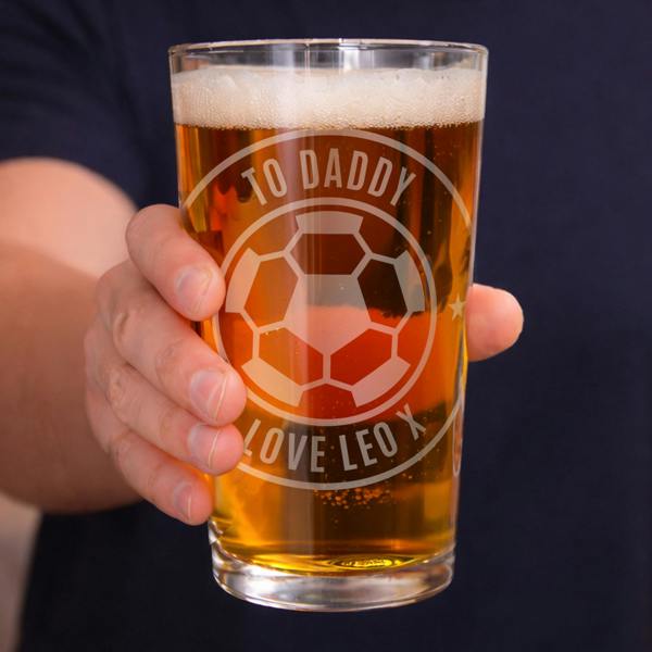 A close-up shot of a man's hand holding a tulip pint glass by Abode Home & Gifts filled with amber beer and foam. The glass is custom engraved with a football badge design reading "TO DADDY" along the top curve and "LOVE LEO X" along the bottom curve.