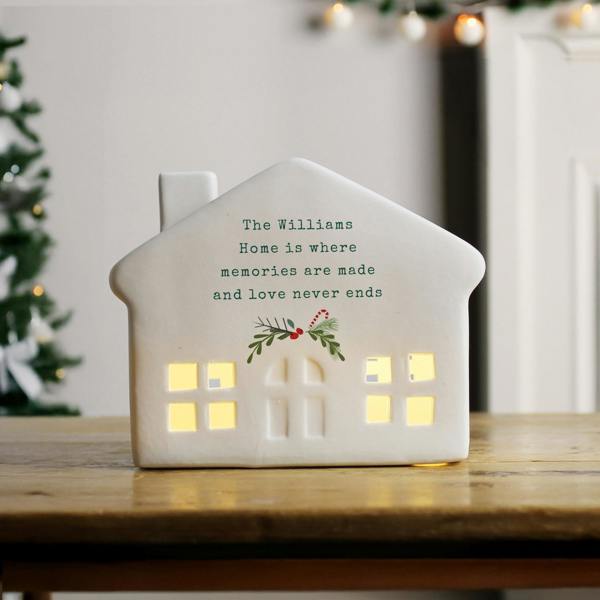 A personalised white ceramic LED light-up house printed with 'The Williams Home is where memories are made' sitting on a wooden table with a Christmas tree in the background.