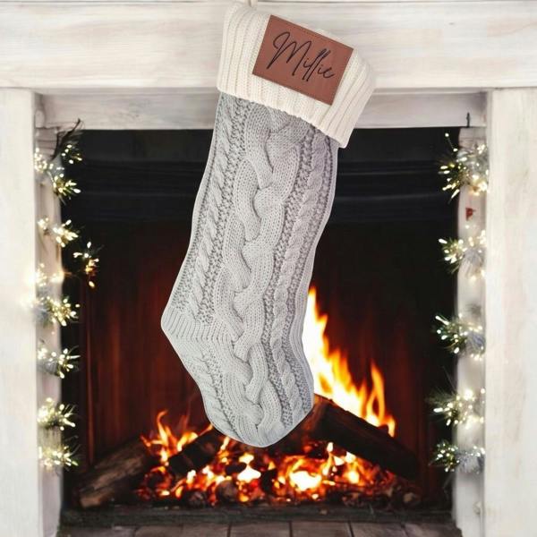 Personalised grey cable-knit Christmas stocking with an engraved faux leather patch reading Millie hanging in front of a glowing fireplace hearth.