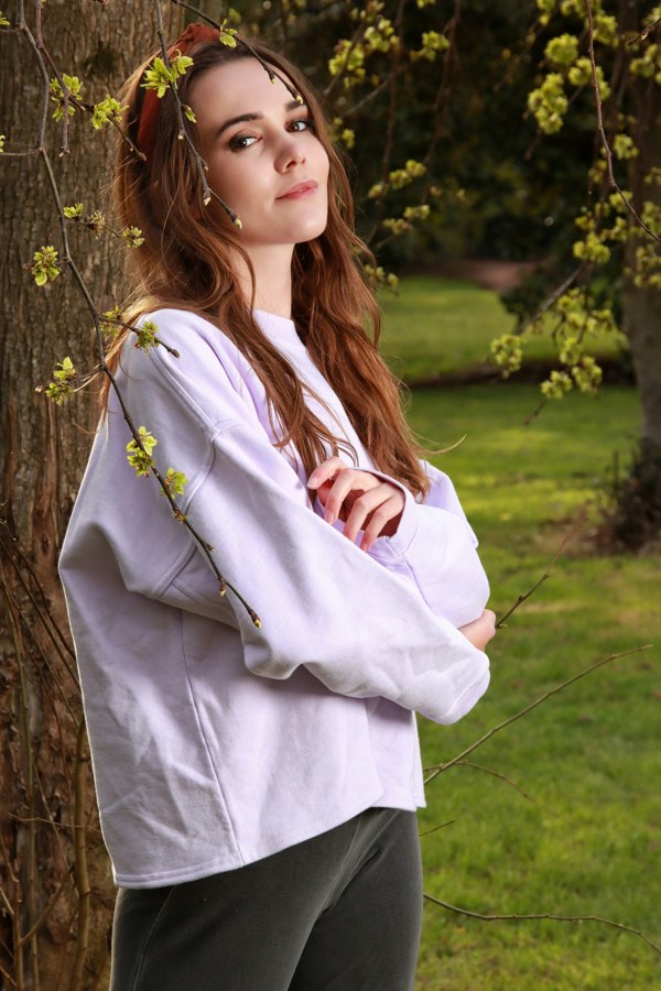 This is a young woman looking at the camera. She is standing in front of a tree in a park which has yellow leaves. She has long hair with a red band. Her arms are loosely folded. She is wearing a bella oversized cropped sweat shirt in lilac. Her tousers are grey.