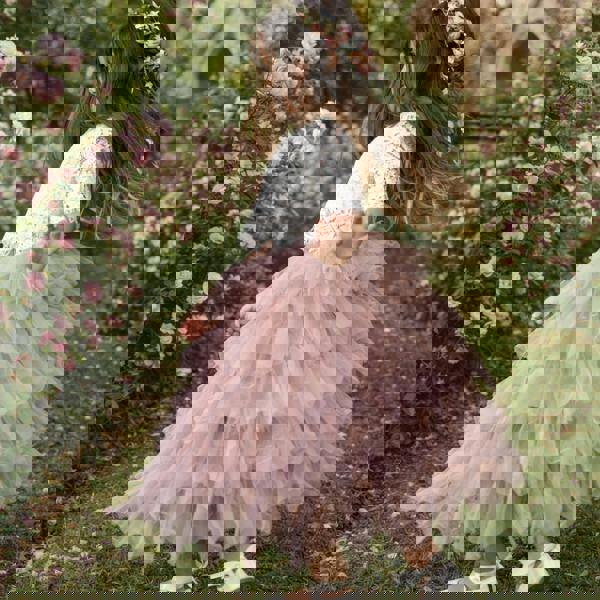 Young girl in a pink dress and white sweater standing in a garden with flowers and a small building in the background.