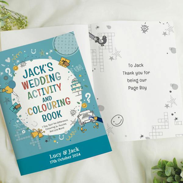 A personalised blue wedding activity and colouring book sits open on a crisp white surface. The cover reads Jack's Wedding Activity and Colouring Book, with a bottom banner printed with Lucy & Jack, 17th October 2024. The open page reveals a custom message layout reading To Jack, Thank you for being our Page Boy.