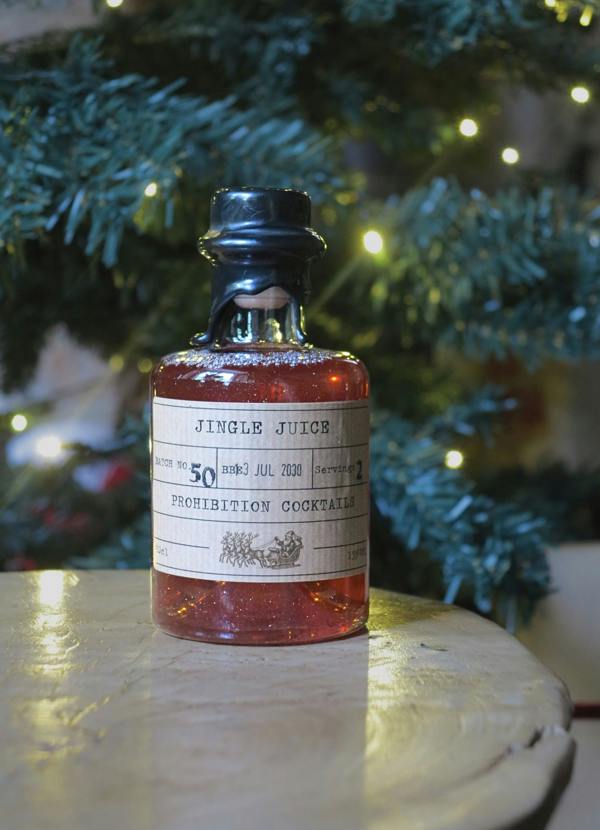 A small glass bottle filled with amber liquid sits on a light wooden surface, with a blurred Christmas tree and warm string lights in the background. The bottle has a black cap and a vintage-style paper label reading 'JINGLE JUICE' and 'PROHIBITION COCKTAILS.