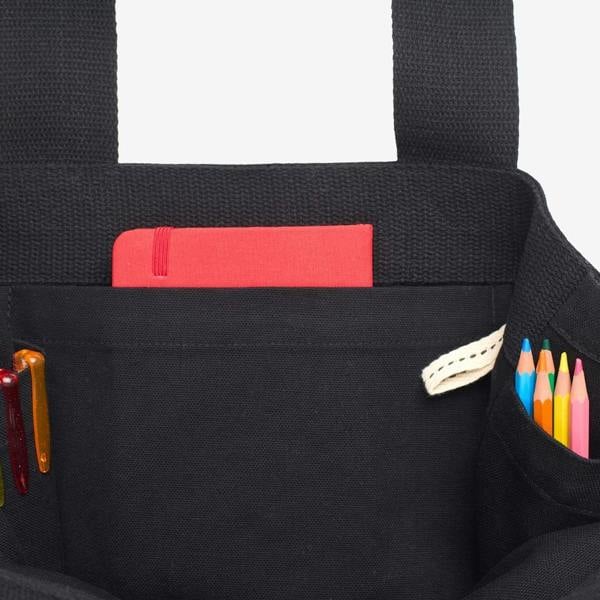 Interior detail view of the black organic cotton Emotional Baggage tote bag showing internal slip pockets containing a red notebook, pens, and colouring pencils.
