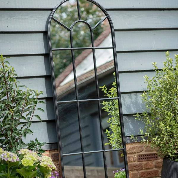 A tall black metal arched window pane mirror mounted on a grey wooden garden wall next to green plants.