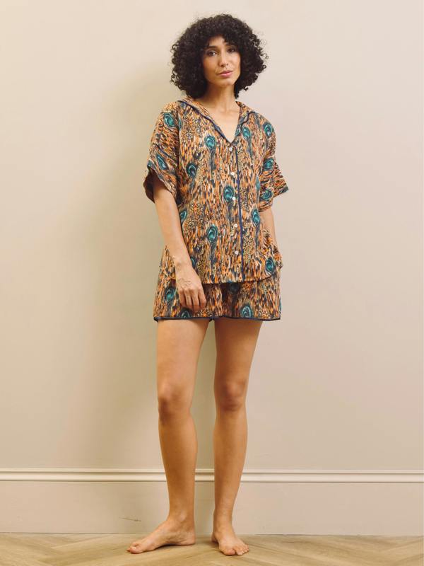 A woman modelling a matching short-sleeve button-front top and shorts pyjama set with an abstract orange, brown, and teal pattern, standing barefoot against a beige wall.