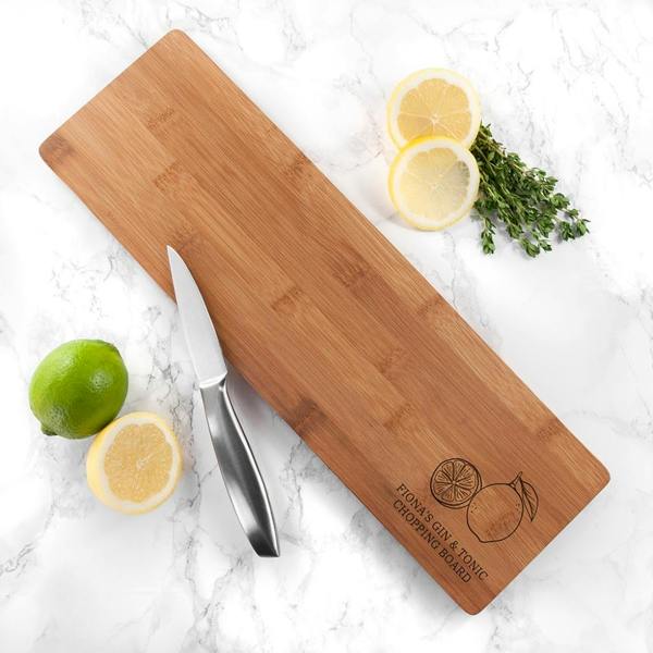A top-down view of a rectangular bamboo Personalised Gin And Tonic Chopping Board engraved with Fiona's Gin & Tonic Chopping Board, resting on white marble with sliced lemons, a lime, and a knife.