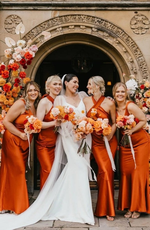 Bride in a white dress with bridesmaids in orange dresses standing in front of an ornate archway.