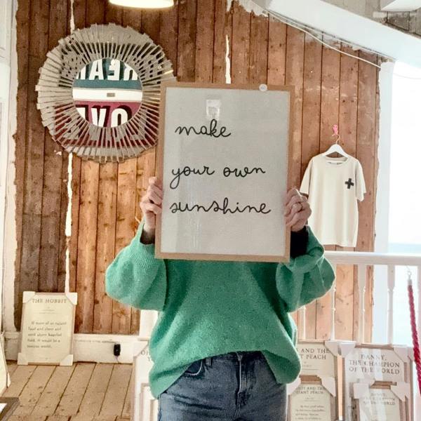 A person in a cosy green jumper holding a solid oak framed white cotton typography print displaying the lowercase handwritten-style quote "make your own sunshine" in front of their face.