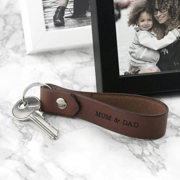 A brown leather loop keyring engraved with the text "MUM & DAD", resting on a marble surface next to a black picture frame featuring a photo of a woman and a child.