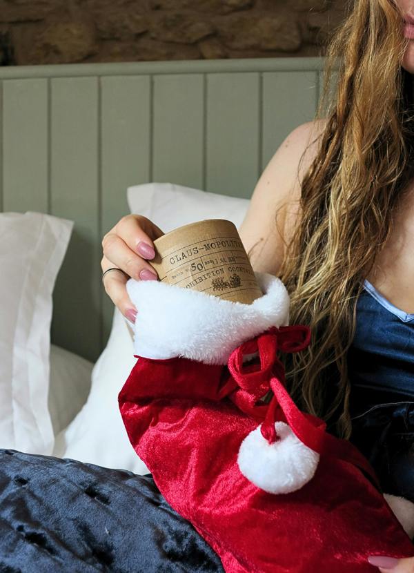 A person sitting in bed holds a red velvet stocking with white faux fur trim and a brown kraft paper label that reads 'Claus-mopolitan by Prohibition Cocktails.'