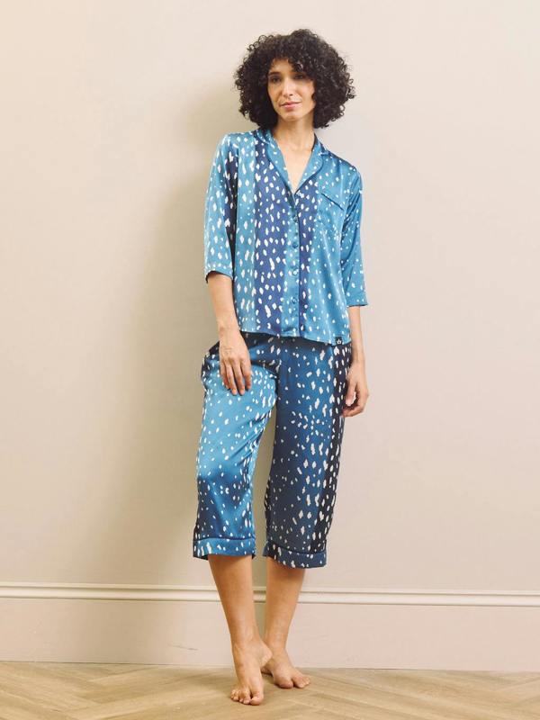 A woman modelling a two-piece capri pyjama set with a blue and white spotted pattern, consisting of a short-sleeve button-up shirt and matching cropped pants, standing against a beige wall.