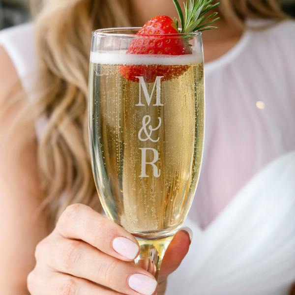 A woman's hand holding up a tall champagne flute by Abode Home & Gifts filled with sparkling wine, garnished with a fresh strawberry and a rosemary sprig. The front of the glass is custom engraved with the capital letters "M", a fixed "&", and an "R" stacked vertically.