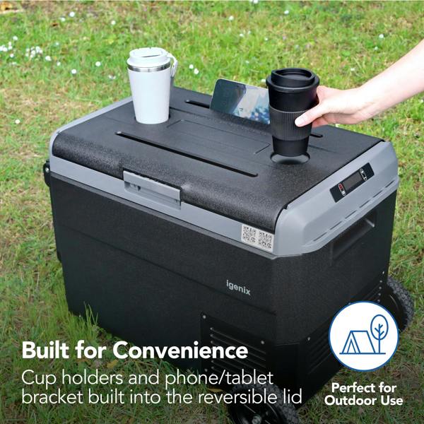 The Igenix 50L Portable Fridge Freezer in black sits on grass, featuring cup holders and a phone/tablet bracket on its lid—shown holding two travel cups and a phone. Text: Built for Convenience and Perfect for Outdoor Use.