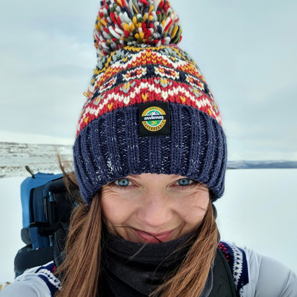 Woman in a snowy landscape is wearing a chunky fairisle knit Super bobble hat navy, red and mustard design. The hat also has a deep ribbed headband and contains reflective yarns and a luxury sherpa fleece lining.