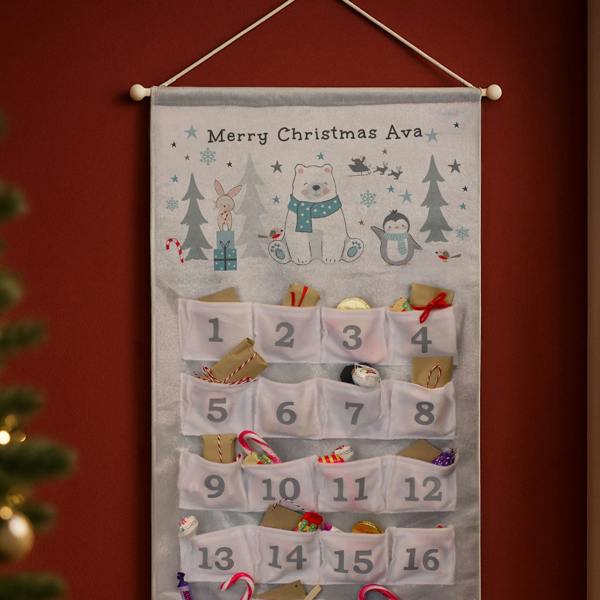 A fabric advent calendar hanging against a dark red wall, featuring a polar bear illustration at the top with the text Merry Christmas Ava, and 24 numbered white pockets filled with small gifts, chocolates, and candy canes.