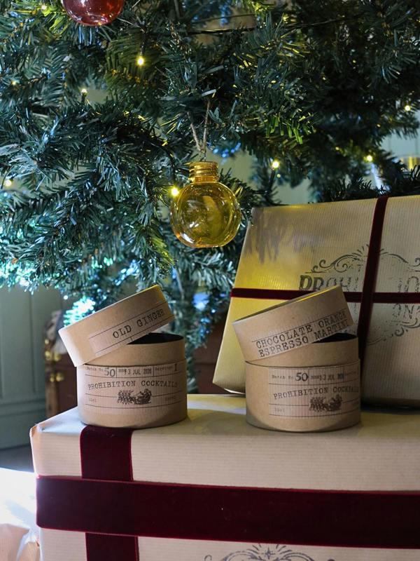 Two gift boxes with lids off, labeled 'Old Ginger' and 'Chocolate Orange Espresso Martini', arranged on top of a wrapped gift box under a decorated Christmas tree with lights and ornaments.