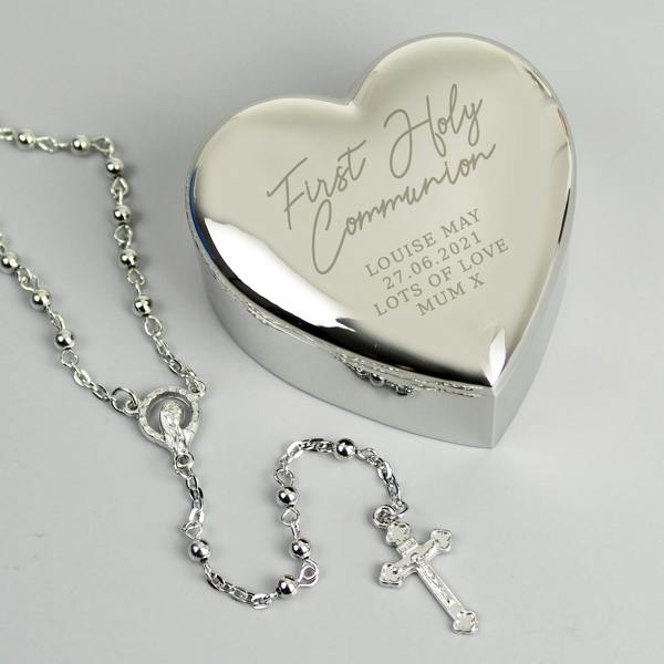 Personalised silver heart-shaped nickel-plated trinket box engraved with 'First Holy Communion' next to a silver rosary bead necklace, presented on a light grey background.