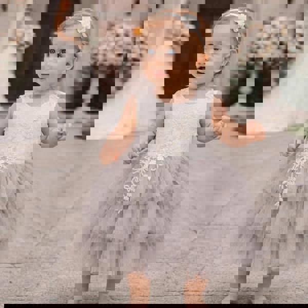 Baby in a gray and white dress with floral details standing outdoors.