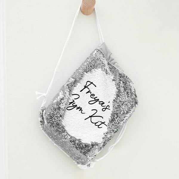 Personalised kids silver sequin drawstring bag hanging on a wooden wall peg, showing the custom revealed text reading Freya's Gym Kit.