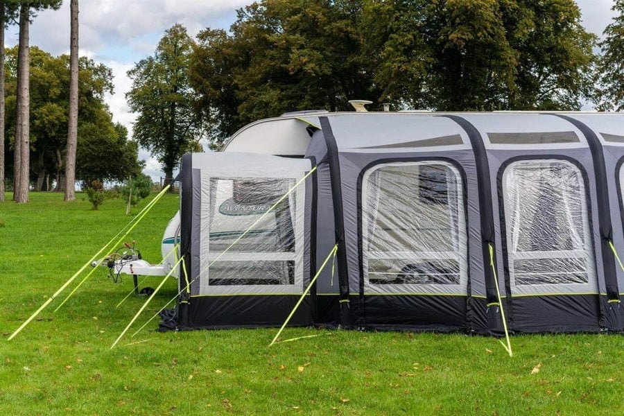 The OLPRO View 420 Caravan Inflatable Porch Awning With Porch Extension attached to a caravan at a campsite with a treeline in the background.