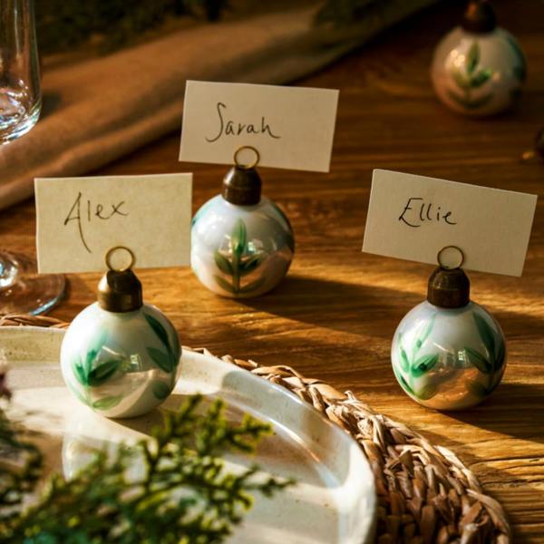 Set of six Bibury hand-painted glass bauble place card holders with handwritten name cards on a wooden Christmas dinner table.