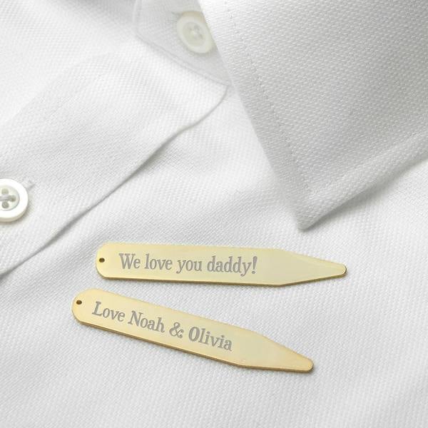 A pair of luxurious 18K gold-plated collar stiffeners lying on a crisp, pristine white dress shirt. The top stiffener is engraved with "We love you daddy!" and the bottom one with "Love Noah & Olivia" in an elegant, classic Serif font.