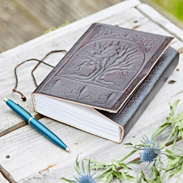 A Tree of Life Leather Journal sits on a wooden table with an embossed cover, next to a blue pen. Partially visible in the foreground are thistle flowers.