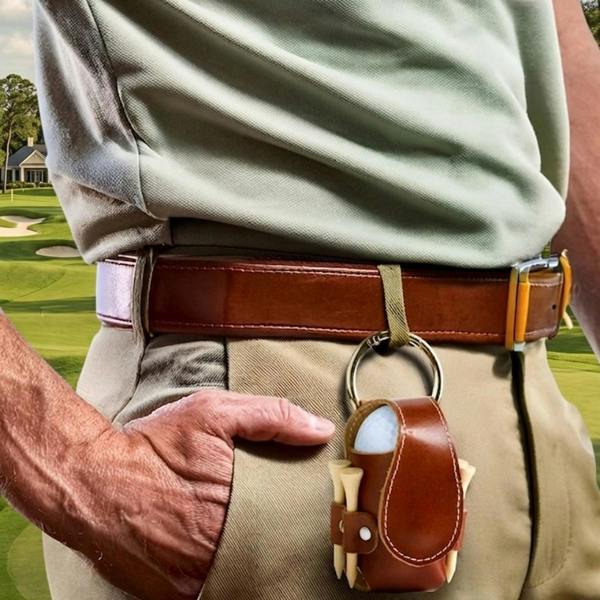 A close-up lifestyle shot of a golfer wearing khaki trousers and a tan leather belt, with a small cognac faux leather caddy case attached. The caddy holds golf balls and wooden tees, set against a blurred green golf course and clubhouse background.