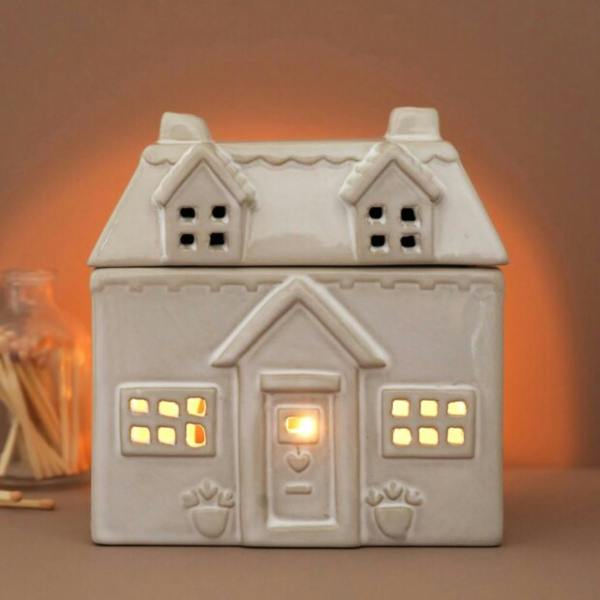 Cream ceramic house wax melt burner glowing from a lit tea light inside on a neutral surface by Abode Home & Gifts