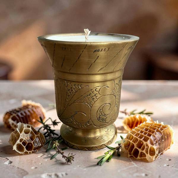 A brass candle holder candle with engraved floral patterns, filled with white wax and a single wick, styled on a stone surface with honeycomb, rosemary sprigs, and a warm, blurred background.