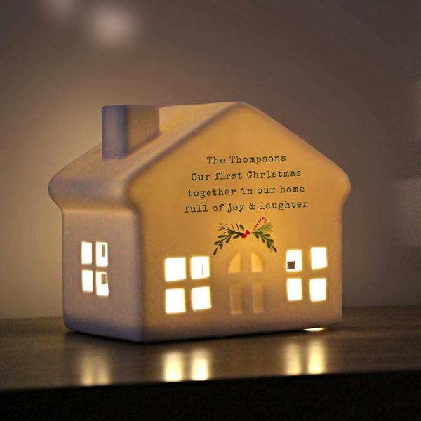 An illuminated personalised white ceramic LED house printed with 'The Thompsons Our first Christmas together in our home full of joy & laughter' glowing in a dimly lit room.