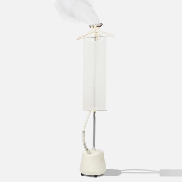A white vertical clothes steamer with a hanger on top, releasing steam. The device has a water tank at the base, a long pole, and a hose attached to the steaming head, all set against a plain light background.