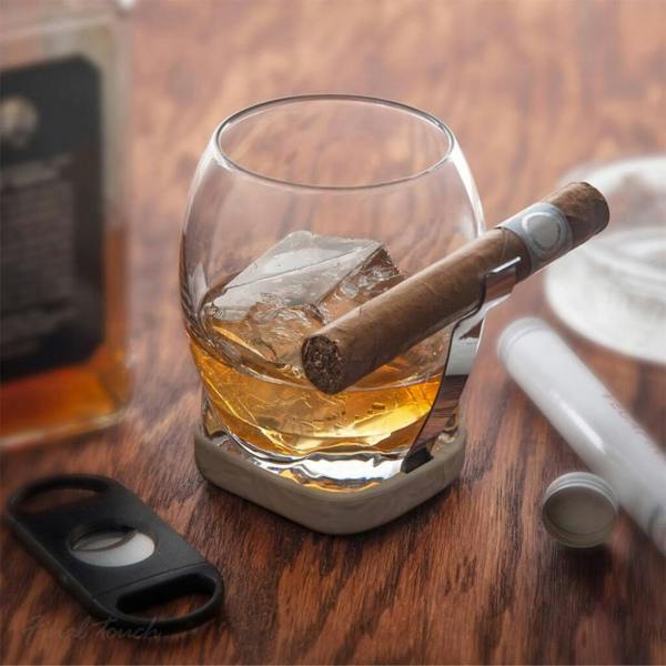 A heavy-bottomed glass whisky tumbler featuring a built-in metal cigar grip holding a cigar safely above a pour of amber spirit.