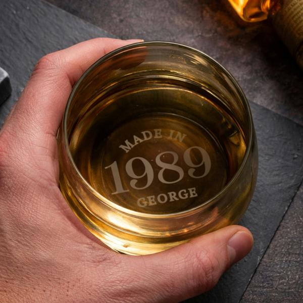 A hand holds a heavy-bottomed glass tumbler filled with an amber-coloured spirit, viewed from directly above. The clear glass base is custom-engraved with the words 'MADE IN 1989 GEORGE' in fixed uppercase lettering. The glass rests over a textured dark slate placemat.