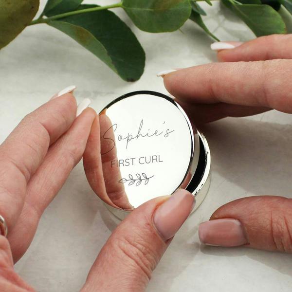 Personalised round silver trinket box being opened by hands, showing the engraving "Sophie's FIRST CURL" with a botanical leaf sprig.