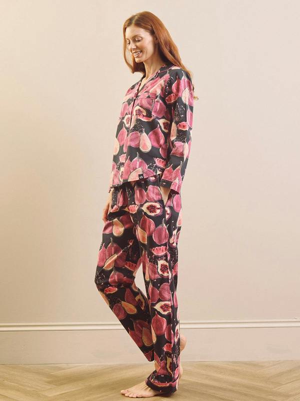 a model wearing a matching pyjama set with a pink and red fig pattern on a black background, set against a neutral beige wall and light wood floor.