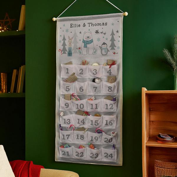 Alt Text: A fabric advent calendar hanging on a deep green wall between a bookshelf and a wooden unit, featuring a polar bear illustration at the top with the text Ellie & Thomas, and 24 numbered white pockets filled with festive sweets, chocolates, and treats.